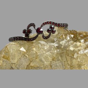 Main photo of antique garnet bracelet displayed on a rock of natural river quartz with the bracelet front forward facing. The largest garnet is to the centre with the band of the bracelet extending from the top right and bottom left like a wave through the central stone and 'ribbon tips' extending out from top left and bottom right of centre stone like the ends of a ribbon. At each end of the 'ribbon ends' are 3 medium sized garnets. The ribbon ends are attached to the main band of the bracelet not loose.