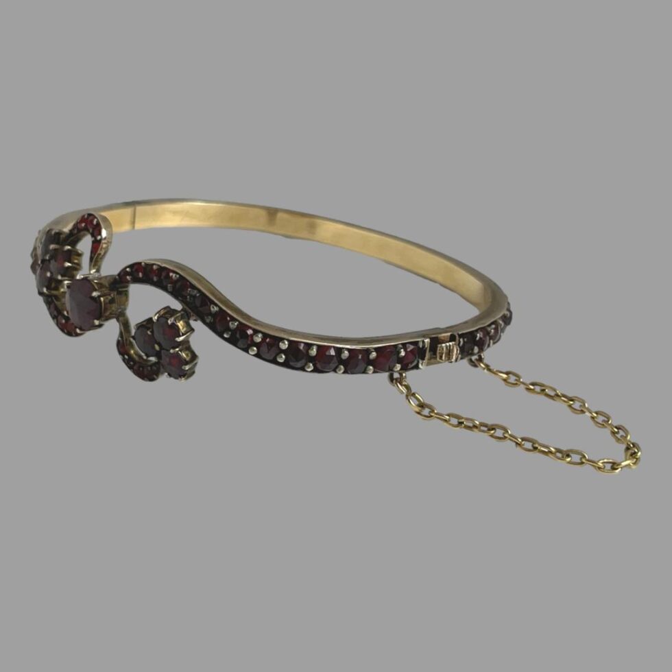 Photo of antique garnet bracelet displayed on a flat surface with the front facing the bottom left corner of photo. The clasp area is at the bottom right and shown closed with the safety chain extending out.