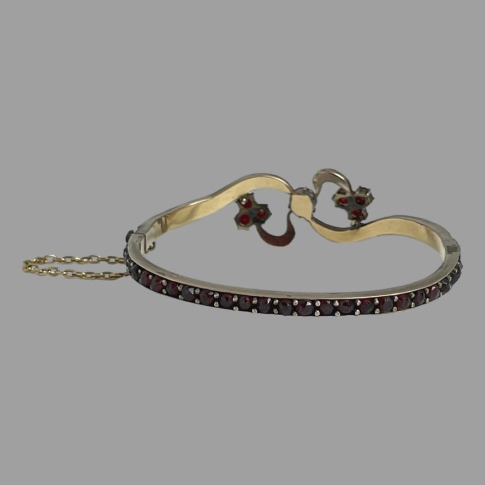 Photo of bracelet displayed on a flat surface with the front facing away from shot and showing the back setting of the front design. The clasp is closed and the safety chain is visible to the left.