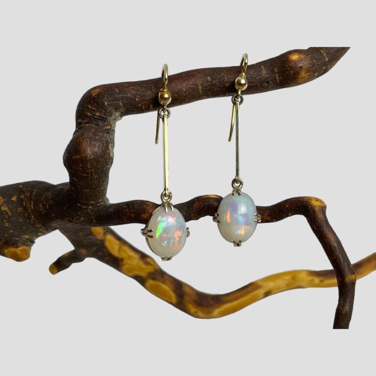 Main photo of antique gold opal earrings displayed hanging side by side from a natural twig of the cherry tree. The earrings are forward facing with incredible colour play emanating from the opals. The earrings are hook fastening with a short straight gold bar attached to the hook and the opals, set with 2 claws at top, bottom, left & right for a secure hold.