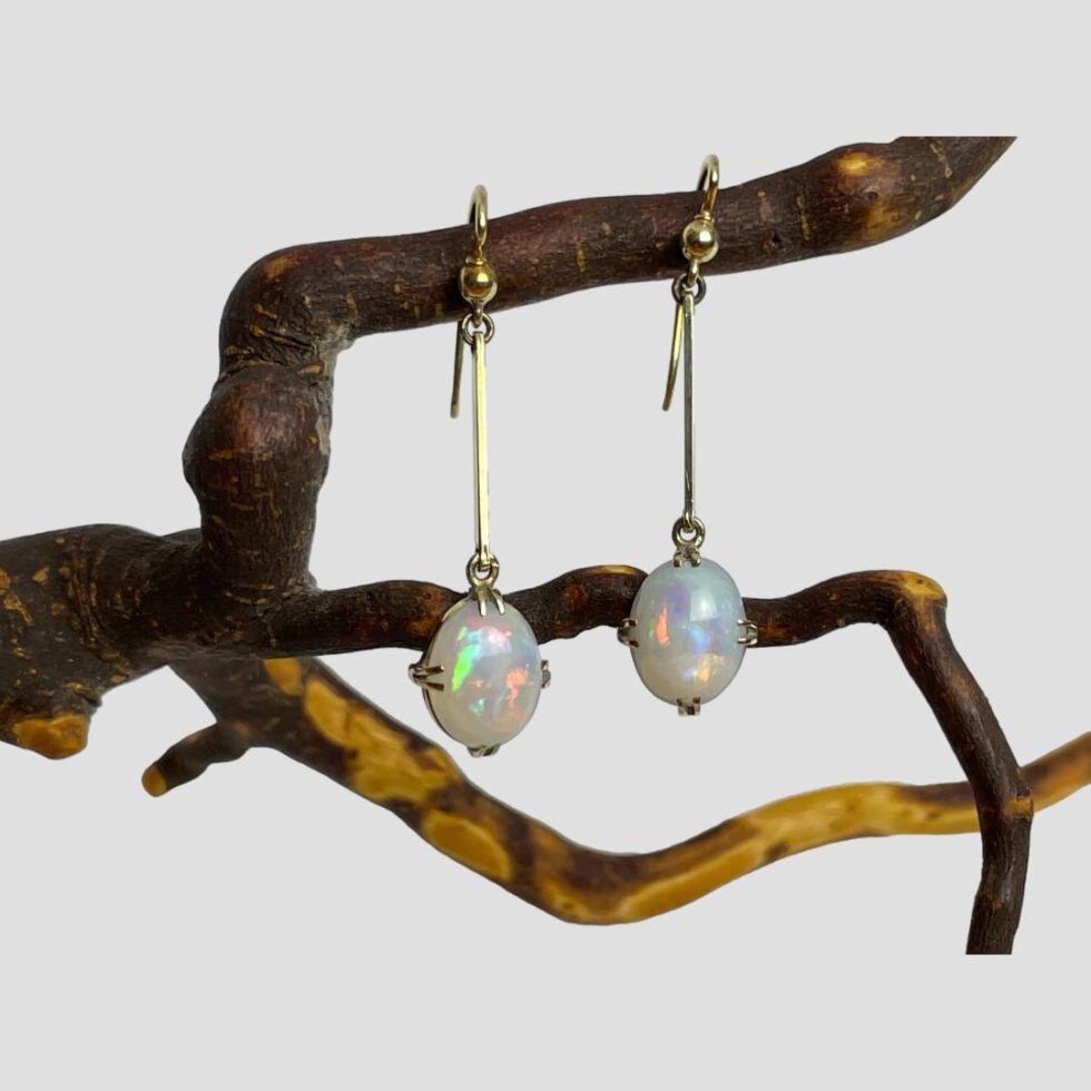 Main photo of antique gold opal earrings displayed hanging side by side from a natural twig of the cherry tree. The earrings are forward facing with incredible colour play emanating from the opals. The earrings are hook fastening with a short straight gold bar attached to the hook and the opals, set with 2 claws at top, bottom, left & right for a secure hold.