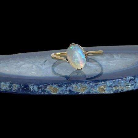 Main photo of 9kt gold opal ring displayed on top of a blue agate slice with the ring front forward facing. Even displayed on a flat surface, the ring front is leaning to the left and the shoulders look as though left one is lower than the other. On close inspection, there are no signs of damage or repair hence probability of it being a design feature. The opal is stunning with intense colour play with flashes of neon turquoise, pinks, oranges and greens.
