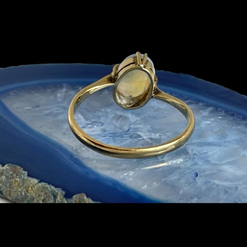 Photo of gold opal ring still displayed on a slice of blue agate and showing the ring from the back. The setting for the opal is visible here and the uneven setting of the band.