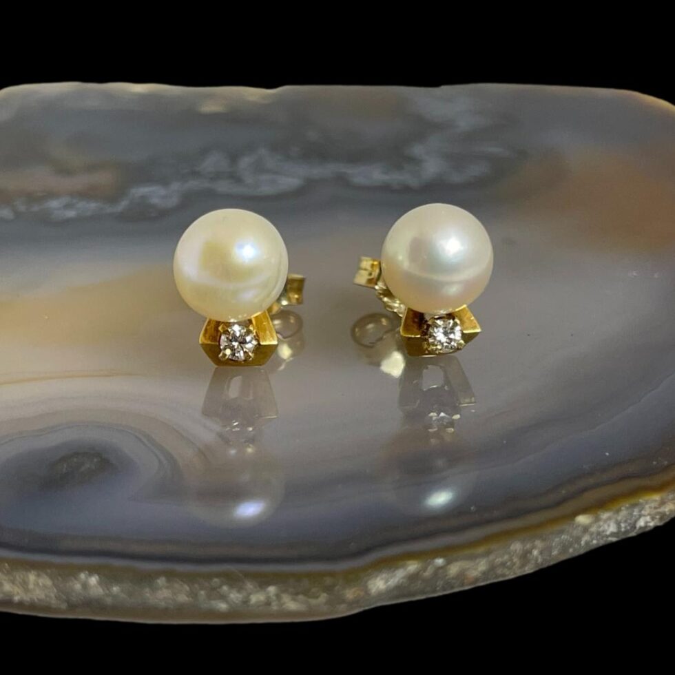 Main photo of gold pearl diamond studs displayed on a slice of natural agate and shown with the earrings fronts forward facing. The earrings are shown free standing with the diamond below the pearls. The diamonds are set within a gold frame and held in place with hook claws. The pearls are a generous size compared to the diamonds but both compliment the other perfectly.