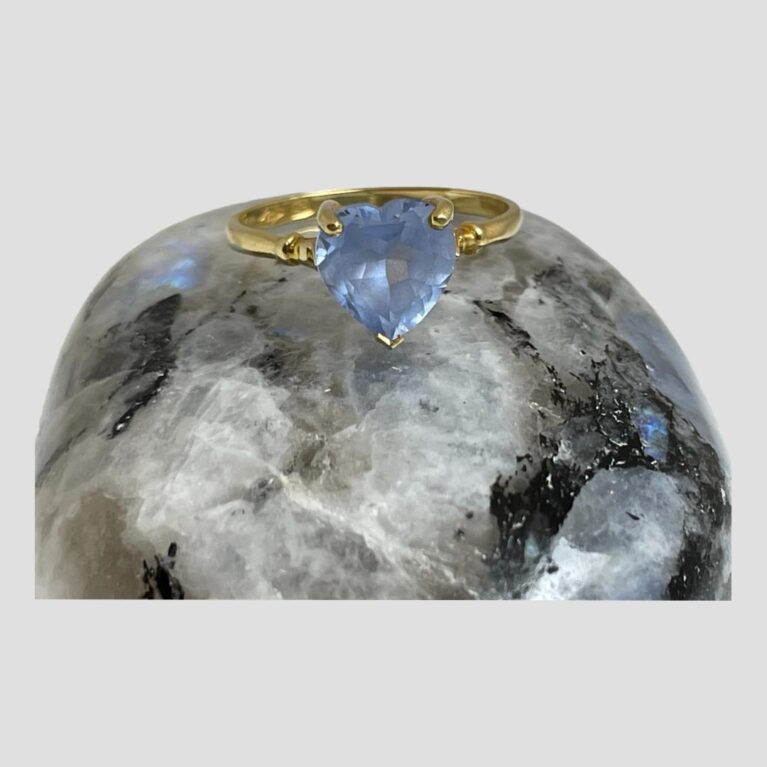 Main photo of gold aquamarine heart ring displayed on top of a smooth rock of natural moonstone with the ring front forward facing. The aquamarine is a a beautiful shade of blue which matches the small flash of blue on the moonstone rock. Simple yellow gold band and setting with a single ball detail to both shoulders.