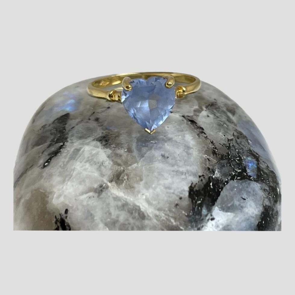Main photo of gold aquamarine heart ring displayed on top of a smooth rock of natural moonstone with the ring front forward facing. The aquamarine is a a beautiful shade of blue which matches the small flash of blue on the moonstone rock. Simple yellow gold band and setting with a single ball detail to both shoulders.
