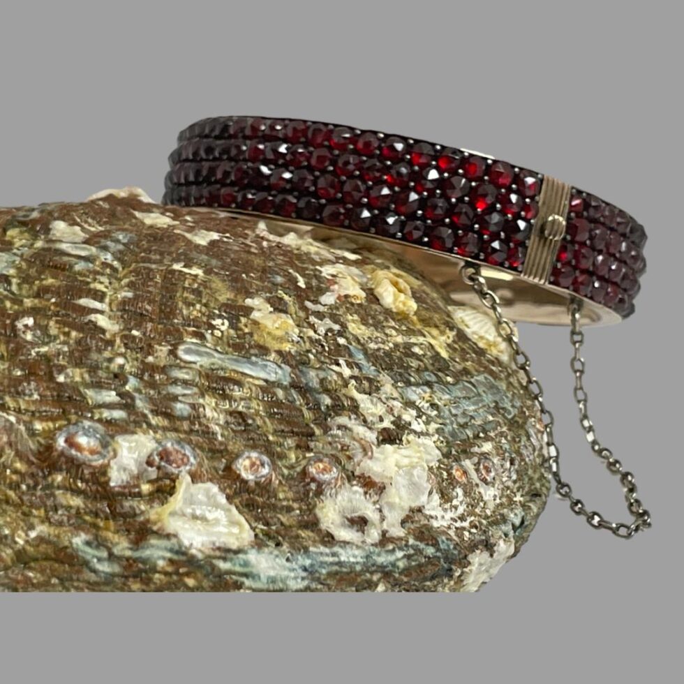 Photo of bracelet this time with one side leaning against the upturned seashell . The clasp area is in the foreground with the safety chain seen hanging in a u shape below. The garnets are a deep red colour in this light.