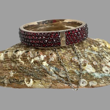 Main photo of Victorian gold garnet bracelet displayed on top of an upturned seashell. The clasp area is in the foreground with safety chain hanging in a u shape below. The facet of each of the garnets is clear and the colour is a rich red. The inside band of the bracelet shows the rose gold colouring which compliments the garnets. The safety chain is a silver colour.
