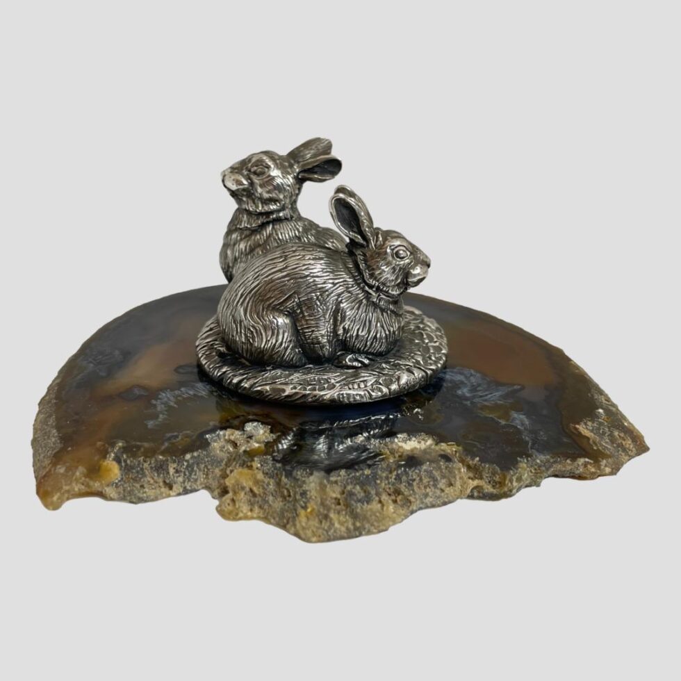 Main photo of of Camelot sterling silver rabbits displayed on top of a slice of natural agate and seen from an eye level. The rabbits are positioned with one in foreground facing the right and one in background facing left. The one at back is on watch duty whilst other is relaxing.