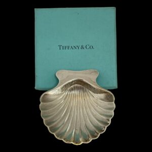 Main photo of Tiffany shell pin dish displayed with back end of scallop edge leaning against the turquoise Tiffany box.; The detailing on the silver scallop shell is quite realistic with a scooped shovel shaped bowl and a smooth back edge. The bowl rests on 2 ball feet at the front and the low dip of the bowl acts as the stabiliser.
