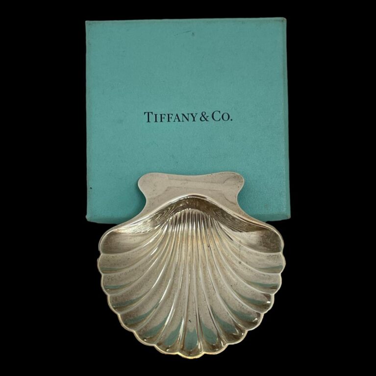 Main photo of Tiffany shell pin dish displayed with back end of scallop edge leaning against the turquoise Tiffany box.; The detailing on the silver scallop shell is quite realistic with a scooped shovel shaped bowl and a smooth back edge. The bowl rests on 2 ball feet at the front and the low dip of the bowl acts as the stabiliser.