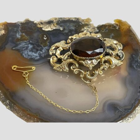 Main photo of Victorian gold filled brooch displayed on top of a slice of natural agate. The photo looks down at the brooch from above with the safety chain shown hooked out to the left in a U shape. This is a very special brooch which would not have looked out of place worn by the flashiest dandies of the day. Lovely frame work around the generous sized stone.