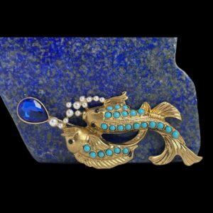 Main photo of vintage fish brooch displayed upright and leaning against a rock of blue lapis giving an underwater feel. The brooch is laid sideways with one fish swimming belly down and other side angled. The lower fish has 3 rows of turquoise in a graduating pattern with longest row running down entire spine to top of n. The side angle fish has one row of turquoise and swims slight above the other with tail fin curving behind its friend. They are both in gilt and appear to be chasing after a royal blue colour teardrop glass and there are tiny pearly beads which represent air bubbles. A truly unique item of quality and beauty.