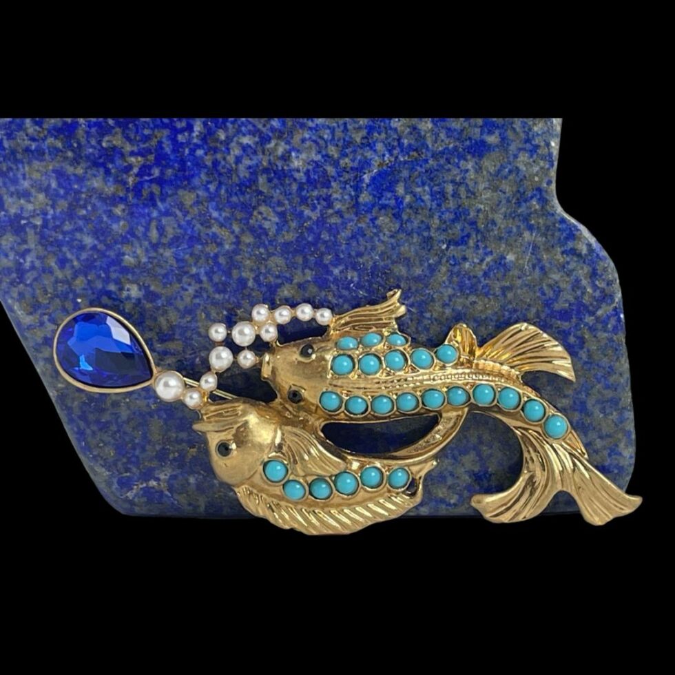 Main photo of vintage fish brooch displayed upright and leaning against a rock of blue lapis giving an underwater feel. The brooch is laid sideways with one fish swimming belly down and other side angled. The lower fish has 3 rows of turquoise in a graduating pattern with longest row running down entire spine to top of n. The side angle fish has one row of turquoise and swims slight above the other with tail fin curving behind its friend. They are both in gilt and appear to be chasing after a royal blue colour teardrop glass and there are tiny pearly beads which represent air bubbles. A truly unique item of quality and beauty.