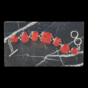 Main photo of red jasper silver bracelet displayed on top of a block of black marble with white inclusions. The bracelet is laid horizontally across in a wave across the marble with the toggle end of clasp on the left and ring end of clasp on the right. The different shapes of the red jasper gives a quirky touch to this chunky bracelet.