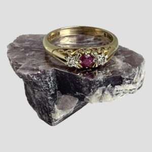 Main photo of gold ruby diamond ring displayed on a rock of natural lepidolite. The ring front is forward facing with the pink ruby contrasting well with the milky purple hue of the lepidolite. The angle of the photo is slightly raised looking diagonally downward showing the front of ring and the inside of back band. The pink round cut ruby is a deep cerise colour and the small diamonds either side are set in white gold making the diamonds appear larger. The gallery is quite intricate in its detailing with the shoulders being very plain in contrast. A very sweet classic look ring.
