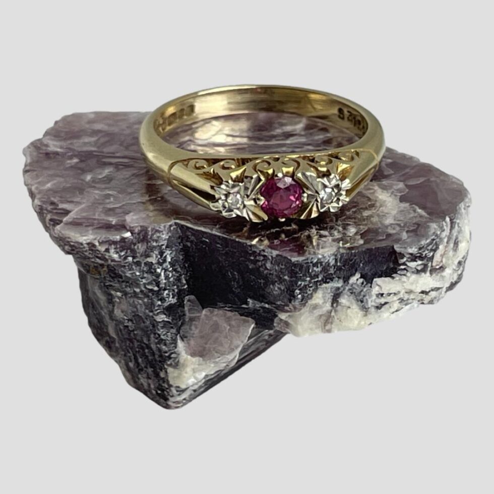 Main photo of gold ruby diamond ring displayed on a rock of natural lepidolite. The ring front is forward facing with the pink ruby contrasting well with the milky purple hue of the lepidolite. The angle of the photo is slightly raised looking diagonally downward showing the front of ring and the inside of back band. The pink round cut ruby is a deep cerise colour and the small diamonds either side are set in white gold making the diamonds appear larger. The gallery is quite intricate in its detailing with the shoulders being very plain in contrast. A very sweet classic look ring.