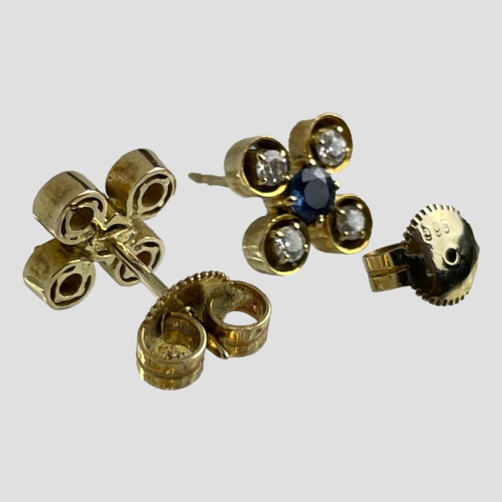 Photo of white & blue sapphire earrings against a plain light grey background with earring in the left foreground facing away from shot displaying the post with butterfly in place. The other earring is facing forward to the right with butterfly removed and placed in front showing the 585 hallmark.