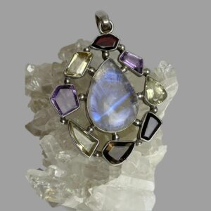 Main photo of silver multi-gem pendant displayed on top of a rock of quartz crystal and the pendant front forward facing. The teardrop shaped large moonstone at the centre is flashing streaks of beautiful blue and with the other stones laid around it like a frame. The different stone shapes adds that extra special feature to this statement pendant with the rainbow moonstone taking centre stage as it should.