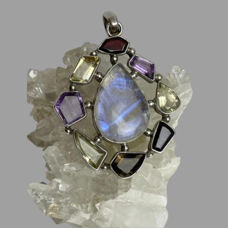 Main photo of silver multi-gem pendant displayed on top of a rock of quartz crystal and the pendant front forward facing. The teardrop shaped large moonstone at the centre is flashing streaks of beautiful blue and with the other stones laid around it like a frame. The different stone shapes adds that extra special feature to this statement pendant with the rainbow moonstone taking centre stage as it should.