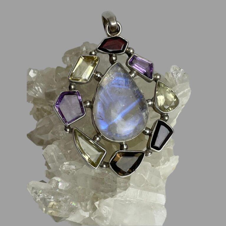 Main photo of silver multi-gem pendant displayed on top of a rock of quartz crystal and the pendant front forward facing. The teardrop shaped large moonstone at the centre is flashing streaks of beautiful blue and with the other stones laid around it like a frame. The different stone shapes adds that extra special feature to this statement pendant with the rainbow moonstone taking centre stage as it should.