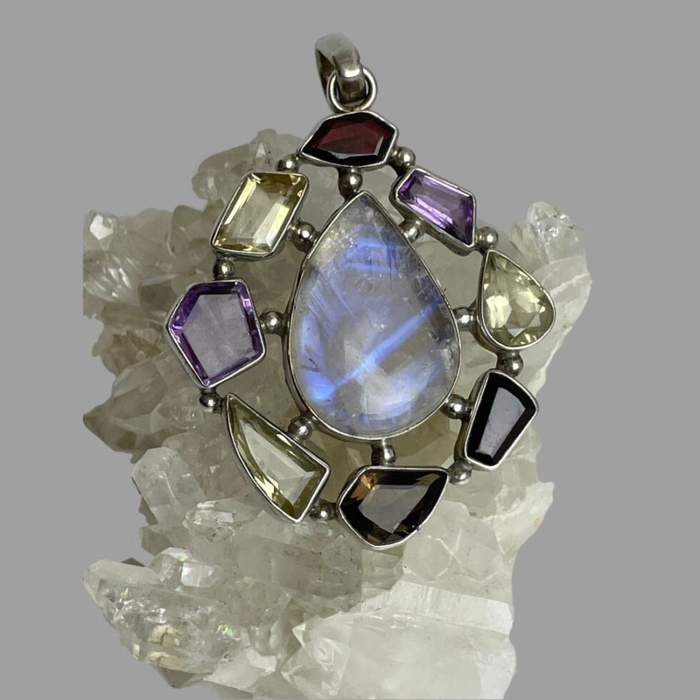 Main photo of silver multi-gem pendant displayed on top of a rock of quartz crystal and the pendant front forward facing. The teardrop shaped large moonstone at the centre is flashing streaks of beautiful blue and with the other stones laid around it like a frame. The different stone shapes adds that extra special feature to this statement pendant with the rainbow moonstone taking centre stage as it should.