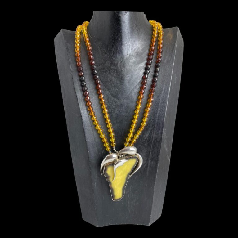 Main photo of vintage multi amber necklace displayed on a black wooden display stand. The large egg yolk amber slice pendant steals the show with its incredible size and colouring-a very yellow egg yolk! Framed in silver with abstract leaf like additions also in silver at the top-almost gives it a fruit like image. The amber beads of the necklace graduate in colour with the darker beads midway down. This is a very striking necklace that will receive many admiring glances!