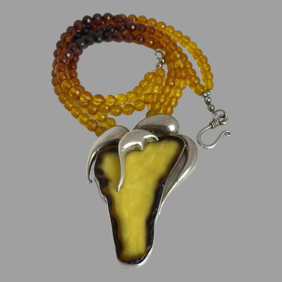 Photo of necklace displayed on a flat surface with a dark grey background. The large pendant is in the lower centre and the faceted bead necklace is laid in a loop above.