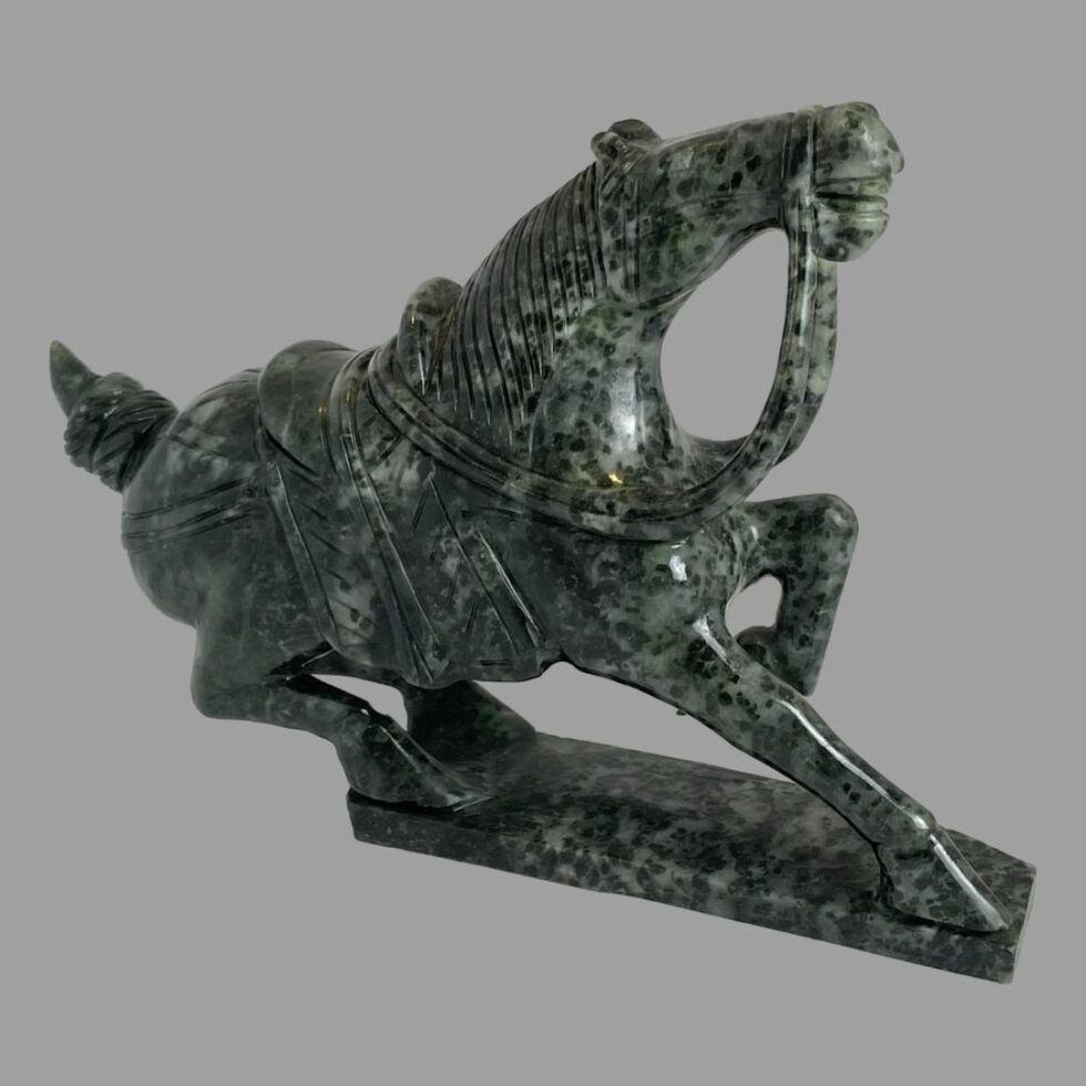 Main photo of serpentine stone warrior horse seen from a side angle with the horse facing to the right. The horse has one front leg raised up, the other straight and its hind legs are both lowered as if ready to pounce any moment. The stone has been well carved with a lot of attention to detail and the mottled stone adds extra character. A perfect gift for someone celebrating the year of the horse!