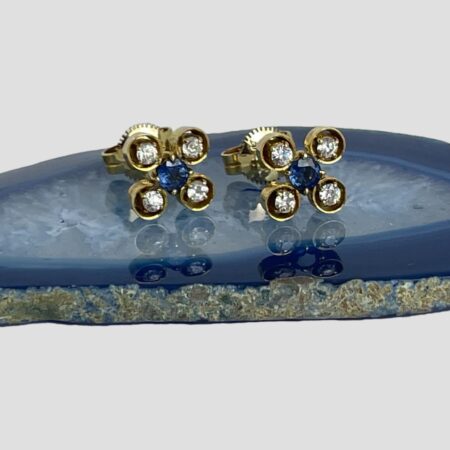 Main pair of white &blue sapphire studs displayed on top of a slice of blue agate with the earring fronts forward facing. The blue sapphire at the centre is held in place by 4 claw hooks. The white or clear sapphires also have claw hooks but each one is also encased in its own gold pocket giving a floral image.