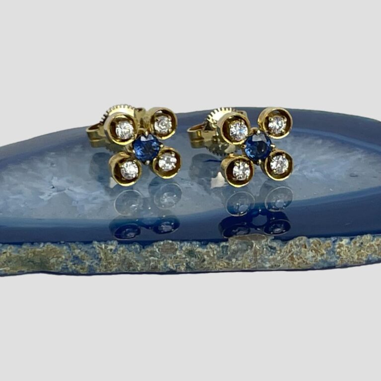 Main pair of white &blue sapphire studs displayed on top of a slice of blue agate with the earring fronts forward facing. The blue sapphire at the centre is held in place by 4 claw hooks. The white or clear sapphires also have claw hooks but each one is also encased in its own gold pocket giving a floral image.