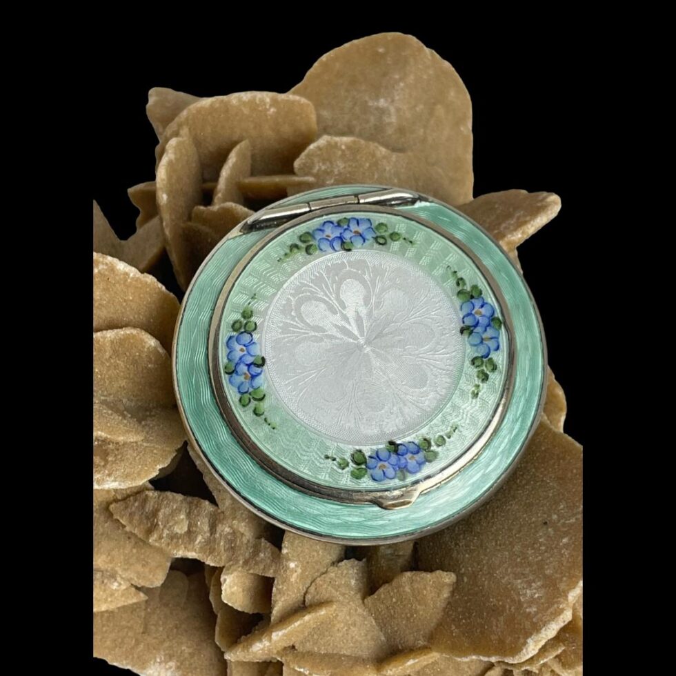 Main photo of 1920s guilloche enamel compact displayed on a rock of natural desert rose with the top of compact on display. The enamel on top is in very good condition with no marks or chips. The main body and outer rim of lid is in minty green with the lid centre having white enamel and blue forget-me-not flowers to 4 sides.