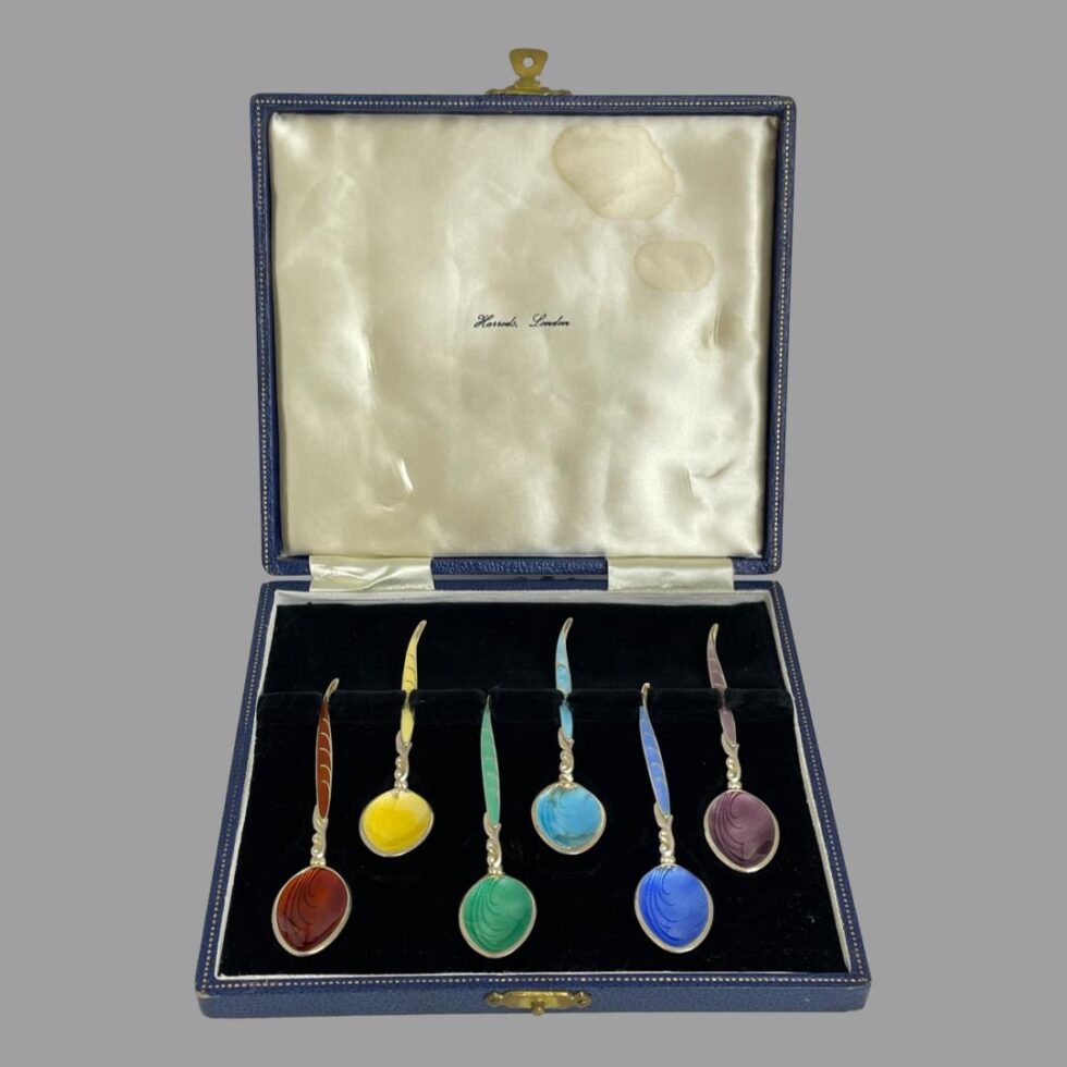 Main photo of silver enamel spoon set displayed with all spoons inside their original fitted case in a rainbow colour formation. Harrods London is printed on the silk inside lid (some staining visible on the silk top right corner)