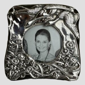 Main photo of London silver photo frame displayed with the front forward facing. This is a very pretty frame with incredible detail. At the top right corner there are 2 geese flying in to land on a lake which is depicted as swirls of water visible to the bottom left corner. All along the lower area of the frame are flowers and reeds. The hallmark is visible to the bottom right corner.