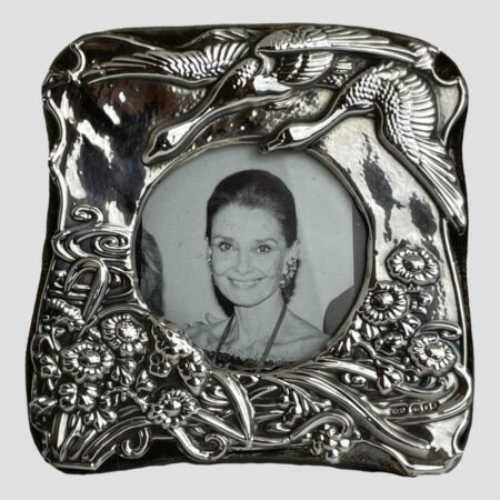 Main photo of London silver photo frame displayed with the front forward facing. This is a very pretty frame with incredible detail. At the top right corner there are 2 geese flying in to land on a lake which is depicted as swirls of water visible to the bottom left corner. All along the lower area of the frame are flowers and reeds. The hallmark is visible to the bottom right corner.
