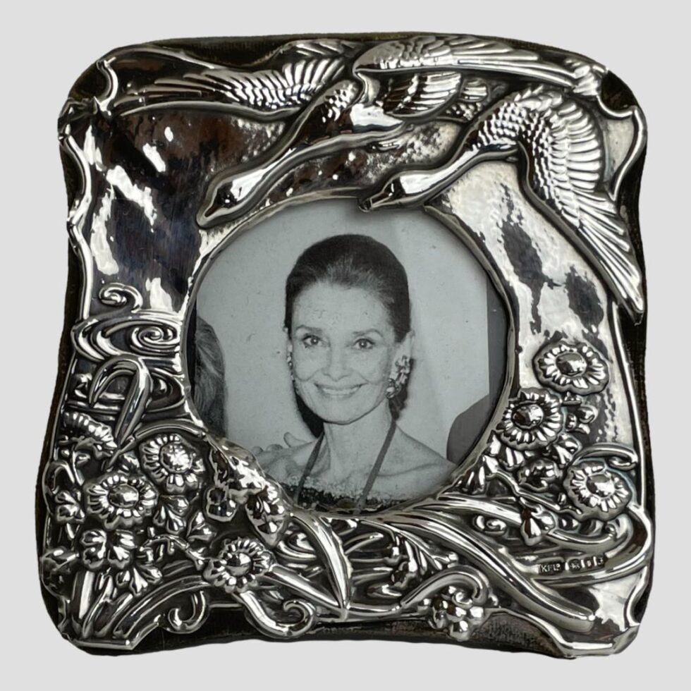 Main photo of London silver photo frame displayed with the front forward facing. This is a very pretty frame with incredible detail. At the top right corner there are 2 geese flying in to land on a lake which is depicted as swirls of water visible to the bottom left corner. All along the lower area of the frame are flowers and reeds. The hallmark is visible to the bottom right corner.