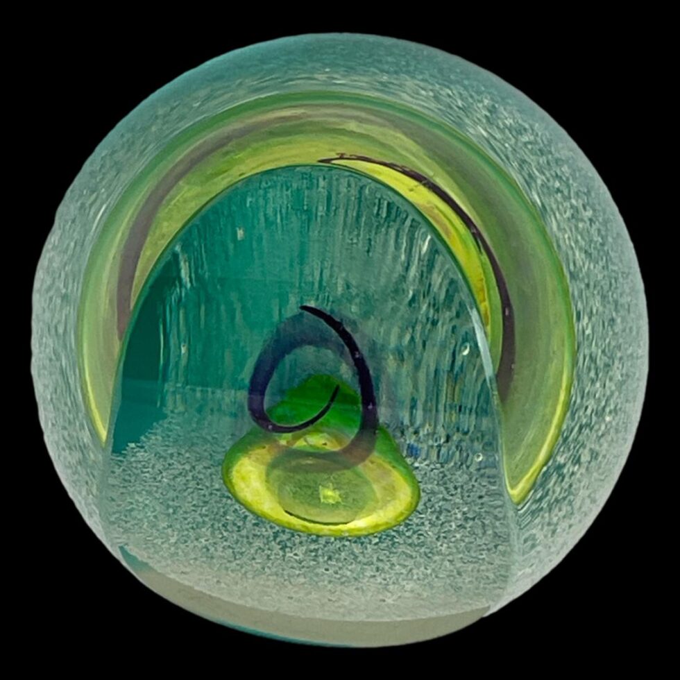Photo looking straight down at Caithness glass druid paperweight from above. From this angle, the grainy white area is almost invisible, the yellow bubble is whole and also reflecting along the top edge and the purple swirl of the druids hat appears as just an open ended circle.