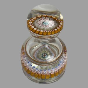 Main photo of c1870 Millefiori glass inkwell seen looking diagonally down from above showing the millefiori detail at the bottom of the lid and bottle. The sides of the millefiori layers around the outer rim are a reddish brown colour with inner columns generally a white. The tips of the columns are where the colour happens with reds, blues and greens. It is difficult to spot any issues with this piece in this photo.