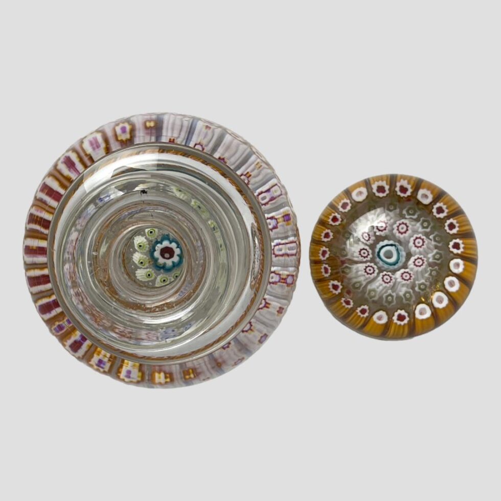 Photo looking straight down at inkwell from above. The lid is removed and placed to the right of the lower bottle. The central millefiori flower is visible through the mouth. There is a small black ink stain on the lip of the bottle which is not visible when the lid is in place.