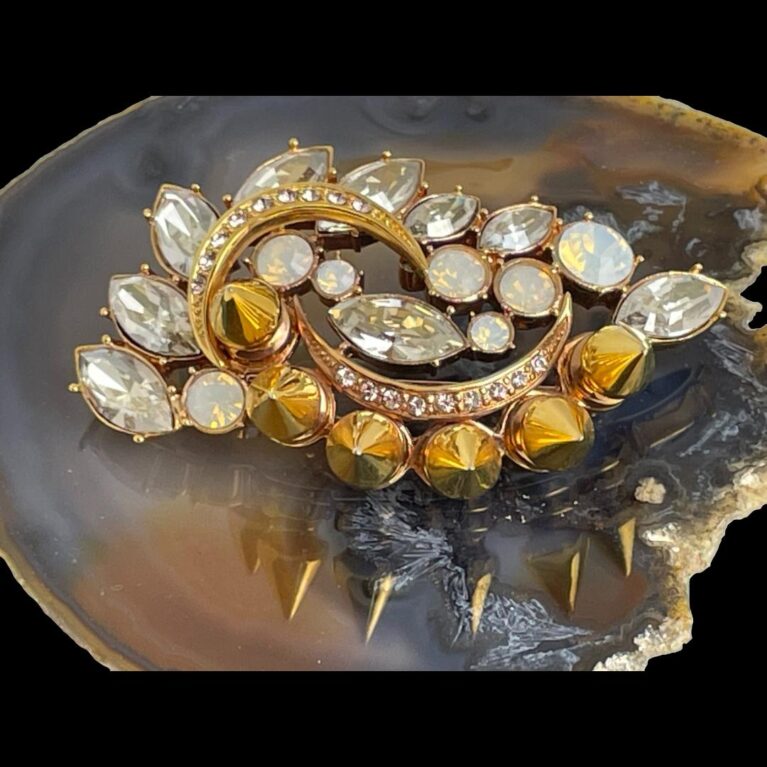 Main photo of Mawi London designer brooch displayed on top of a slice of natural agate. The row of gilt spikes is arched along the bottom in this photo with the crystals leaves scattered around 2 crescent moon shaped details within the brooch. The reflection of the gilt spikes can be seen on the agate. This is a fantastic piece of jewellery!