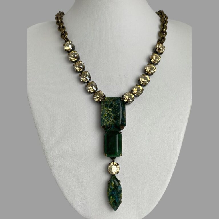 Main photo of Philippe Ferrandis designer necklace displayed on a white coloured display stand with the necklace front forward facing. The long non detachable pendant is a deep green shade like moss agate with a single citrus coloured crystal separating the oval green at bottom from the 2 rectangular green above. 6 citrus coloured crystals line the lower part of the necklace chain to either side followed by a thick brassy coloured chain. The extension chain has a single citrus crystal on the end.