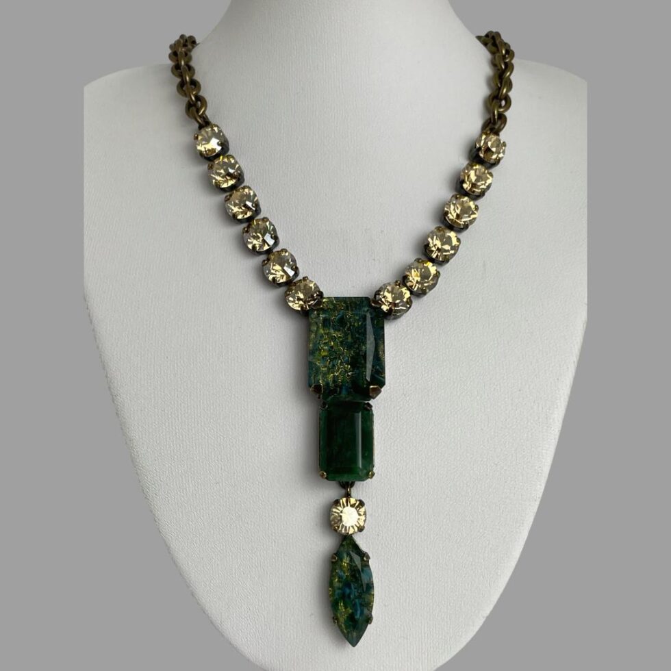 Main photo of Philippe Ferrandis designer necklace displayed on a white coloured display stand with the necklace front forward facing. The long non detachable pendant is a deep green shade like moss agate with a single citrus coloured crystal separating the oval green at bottom from the 2 rectangular green above. 6 citrus coloured crystals line the lower part of the necklace chain to either side followed by a thick brassy coloured chain. The extension chain has a single citrus crystal on the end.