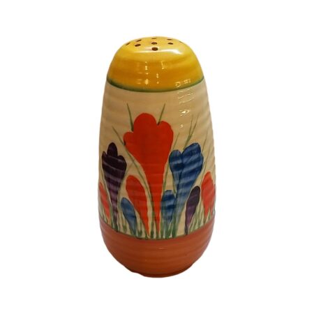 Main photo of Clarice Cliff Lynton sugar sifter seen from a near eye level angle with different coloured crocus flowers in the foreground. The top of shaker is yellow and around the base is orange. Lovely cheerful colour combination!