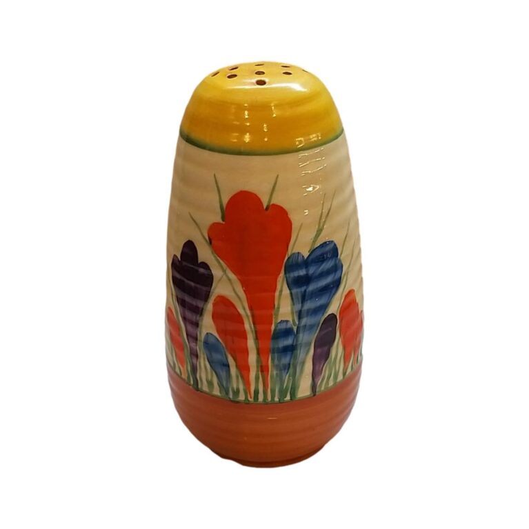Main photo of Clarice Cliff Lynton sugar sifter seen from a near eye level angle with different coloured crocus flowers in the foreground. The top of shaker is yellow and around the base is orange. Lovely cheerful colour combination!