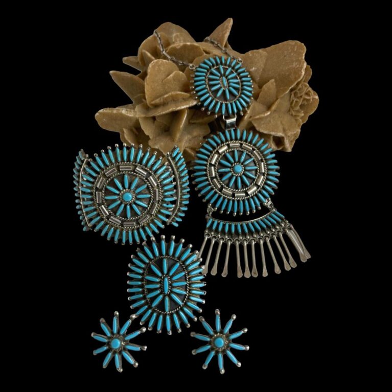 Main photo of signed Zuni needlepoint set showing all 4 pieces displayed against a black background with a rock of natural desert sand rose as prop. The turquoise sections of the necklace is draped on the desert sand rose whilst the cuff, ring then stud earrings are displayed on the flat surface in front of the rock. The sleeping beauty turquoise stones are a stunning blue - one of the highest regarded turquoise stones for their vibrant beauty. An absolutely stunning set that can be worn together or separately as the mood takes you.