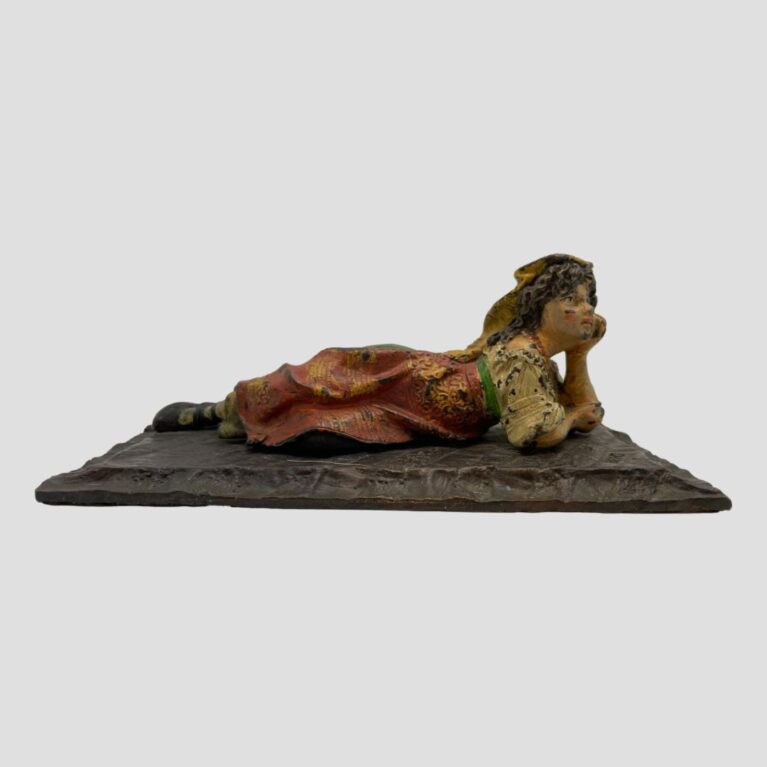 Main photo of bronze Bedouin girl displayed showing her whole reclined length. She lies partially on her side with top end face down and her left cheek cupped in one hand. She looks dreamily out, maybe daydreaming and has a reflective look in her eyes. There is quite some wear to the paint with dark patches of bronze coming through throughout but there is more paint in tact to show the lovely colours used. She is wearing a flat head cover which trails behind in a dark mustard colour, an off the shoulder cream colour blouse, a long green skirt with a red apron at the front. She is also wearing some red necklaces. Her hair is shoulder length, wavy & brown and her eyes are a hazel brown colour. Above all, the dreaminess of this bronze is the magic.