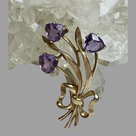 Main photo of gold amethyst tulips brooch displayed leaning against a rock of natural quartz crystal with the amethyst tulip flower heads at the top. The tulips are faceted upside down heart shaped set on a gold backing with 4 teeth holding each stone in place. The rest of the brooch is in gold with 3 stems long leaves and a bow at the stem ends. It has a curved design feature so would look perfect on a lapel or collar. The amethysts are a lovely shade of purple - not too dark and not light!