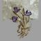Main photo of gold amethyst tulips brooch displayed leaning against a rock of natural quartz crystal with the amethyst tulip flower heads at the top. The tulips are faceted upside down heart shaped set on a gold backing with 4 teeth holding each stone in place. The rest of the brooch is in gold with 3 stems long leaves and a bow at the stem ends. It has a curved design feature so would look perfect on a lapel or collar. The amethysts are a lovely shade of purple - not too dark and not light!