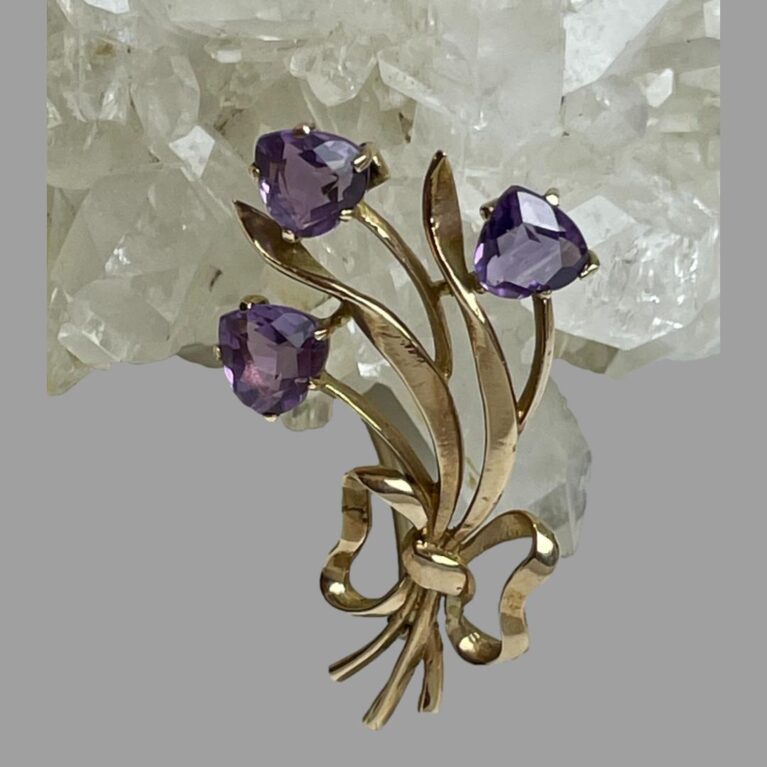 Main photo of gold amethyst tulips brooch displayed leaning against a rock of natural quartz crystal with the amethyst tulip flower heads at the top. The tulips are faceted upside down heart shaped set on a gold backing with 4 teeth holding each stone in place. The rest of the brooch is in gold with 3 stems long leaves and a bow at the stem ends. It has a curved design feature so would look perfect on a lapel or collar. The amethysts are a lovely shade of purple - not too dark and not light!