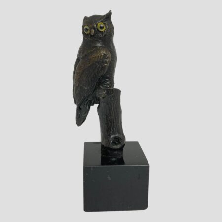 Main photo of vintage small owl bronze. The owl has his head turned to the camera so its yellow glass eyes are looking at you but its breast is facing right with tail hanging down on the left. The branch that it is perched on is not visible from this angle but the trunk can be seen. The wonderful thing about this piece is that the owl is doing what an owl does - turning its head to impossible degrees!